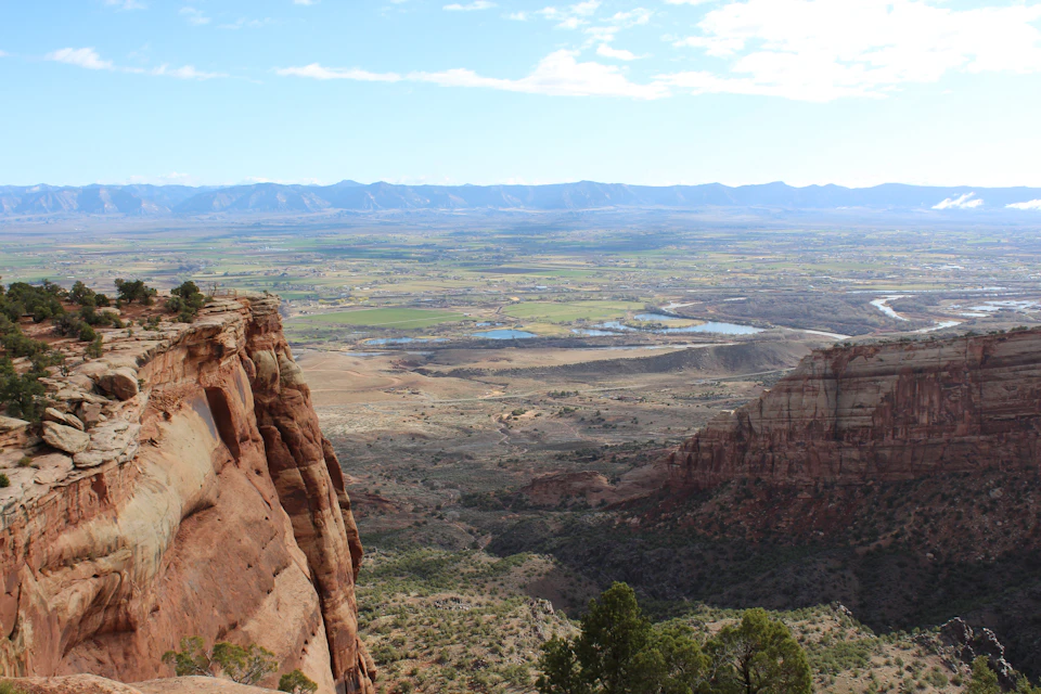 The image depicts a breathtaking view from a high vantage point, overlooking a valley nestled between two towering cliffs. The valley is a vast expanse of green fields and grasslands that stretch out into the distance, with a river meandering through it. The sky above is a clear blue, dotted with fluffy white clouds. On either side of the valley, there are mountains rising up against the horizon, their peaks shrouded in mist. The image is taken from an elevated position, providing a panoramic view of this picturesque landscape.