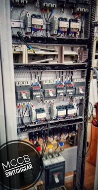 An electrical switchgear panel with multiple circuit breakers and wiring is organized within a metal frame. Colored wires are neatly attached, and labels or indicators can be seen on some components. The environment suggests an industrial or technical setting.