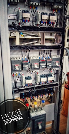 An electrical switchgear panel with multiple circuit breakers and wiring is organized within a metal frame. Colored wires are neatly attached, and labels or indicators can be seen on some components. The environment suggests an industrial or technical setting.