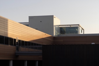 A modern building exterior with a mix of horizontal brown paneling and large glass windows. The architecture features clean lines and geometric shapes, enhanced by the warm light casting shadows on the structure.