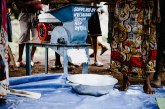 A group of people stands around a blue mechanical grinder with a red motor on a metal table. The blue machine has writing on it, indicating it was supplied by an organization. A colorful fabric with swirling patterns is worn by one of the individuals. The scene takes place outdoors on a dirt surface with a blue plastic sheet on the ground and a metal bowl nearby.