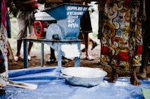 A group of people stands around a blue mechanical grinder with a red motor on a metal table. The blue machine has writing on it, indicating it was supplied by an organization. A colorful fabric with swirling patterns is worn by one of the individuals. The scene takes place outdoors on a dirt surface with a blue plastic sheet on the ground and a metal bowl nearby.