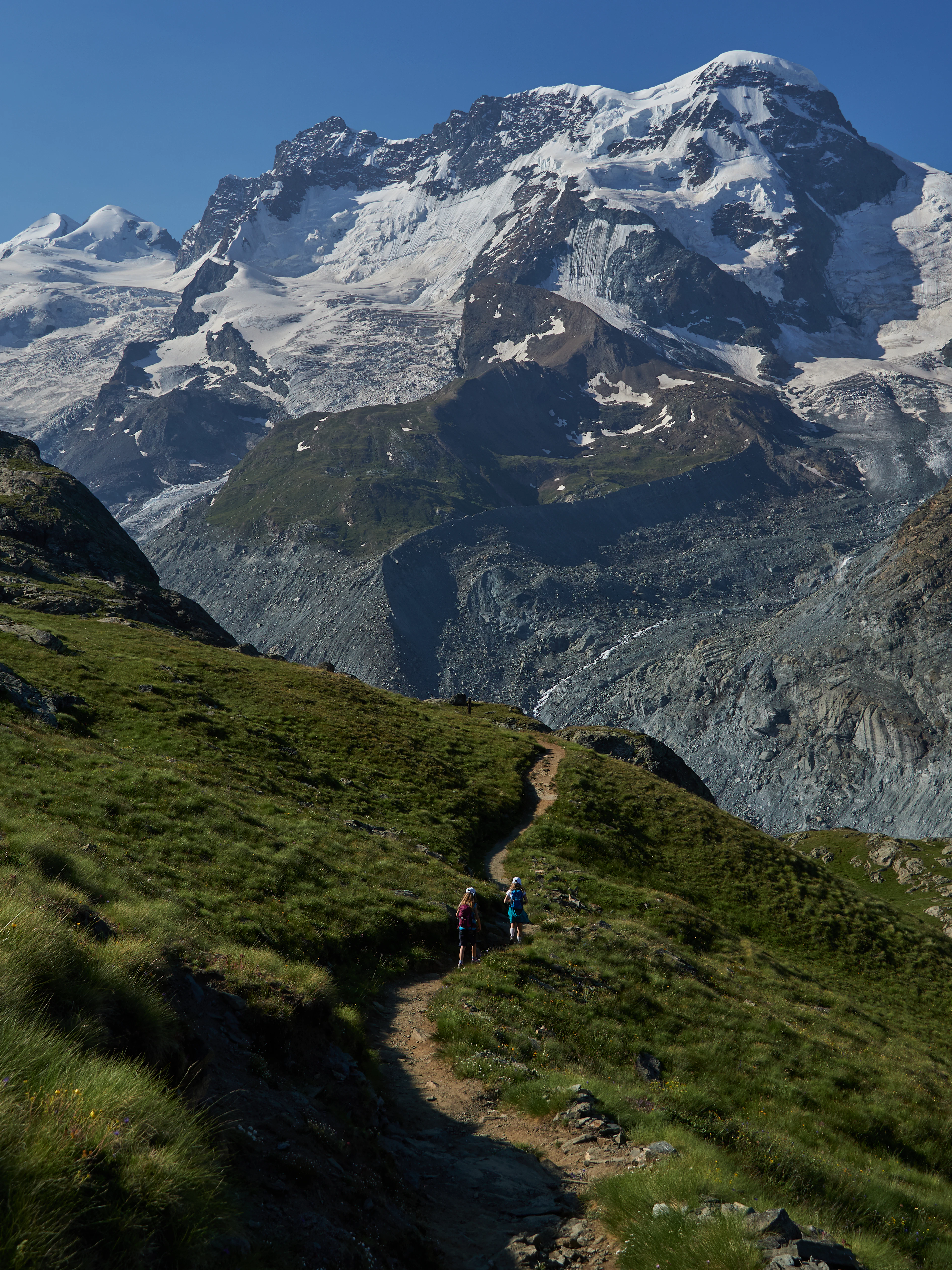 Two hikers follow a ridge trail beneath a massive glaciated mountain wall and icefall.