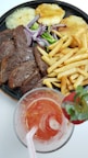 A plate features sliced grilled steak accompanied by golden fries, grilled pineapple, red onion slices, and green vegetables. Beside the plate is a glass filled with a red beverage, garnished with a strawberry.