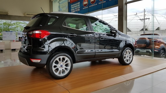 A sleek black SUV is displayed on a shiny wooden platform inside a car dealership with large glass windows showing another vehicle outside.