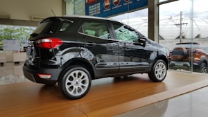 A sleek black SUV is displayed on a shiny wooden platform inside a car dealership with large glass windows showing another vehicle outside.