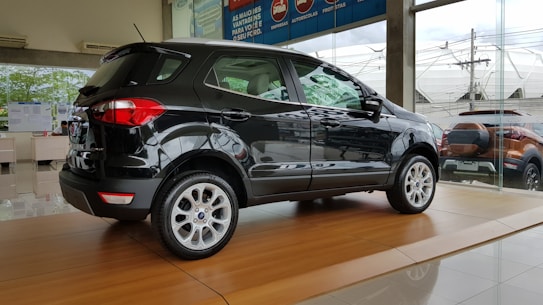 A sleek black SUV is displayed on a shiny wooden platform inside a car dealership with large glass windows showing another vehicle outside.