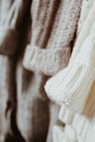 Close-up view of several knitted sweaters in soft, neutral tones hanging together. The textures of the various knit patterns are clearly visible, showcasing different stitch types and detailing.