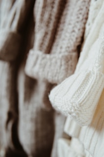 Close-up view of several knitted sweaters in soft, neutral tones hanging together. The textures of the various knit patterns are clearly visible, showcasing different stitch types and detailing.