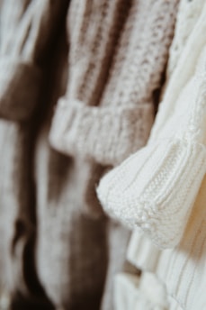 Close-up view of several knitted sweaters in soft, neutral tones hanging together. The textures of the various knit patterns are clearly visible, showcasing different stitch types and detailing.