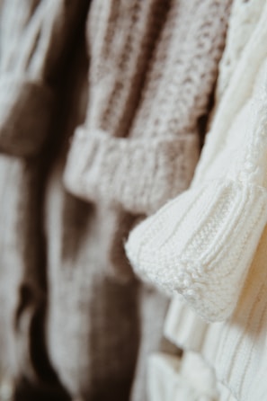 Close-up view of several knitted sweaters in soft, neutral tones hanging together. The textures of the various knit patterns are clearly visible, showcasing different stitch types and detailing.