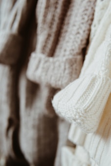 Close-up view of several knitted sweaters in soft, neutral tones hanging together. The textures of the various knit patterns are clearly visible, showcasing different stitch types and detailing.