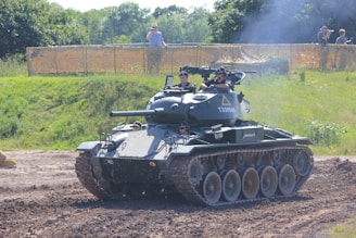 A military tank is maneuvering on a dirt path with several people visible inside the vehicle, wearing uniforms and helmets. The setting is outdoors, with green foliage and trees in the background, and a spectator area fenced off with netting where a few people are watching.