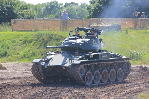 A military tank is maneuvering on a dirt path with several people visible inside the vehicle, wearing uniforms and helmets. The setting is outdoors, with green foliage and trees in the background, and a spectator area fenced off with netting where a few people are watching.