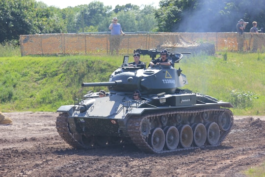 A military tank is maneuvering on a dirt path with several people visible inside the vehicle, wearing uniforms and helmets. The setting is outdoors, with green foliage and trees in the background, and a spectator area fenced off with netting where a few people are watching.