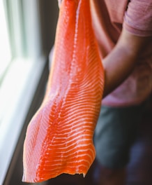 A person is holding a large, fresh piece of salmon fillet with distinct lines of fat running across the bright orange flesh. The setting is indoors, near a window that casts natural light on the salmon.
