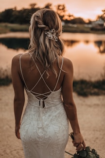 A woman in an elegant, white lace dress stands with her back to the camera, facing a serene, reflective body of water at sunset. Her blonde hair is styled in loose curls and adorned with an ornate hairpiece. She holds a small bouquet of flowers in one hand, complementing the romantic ambiance of the scene.