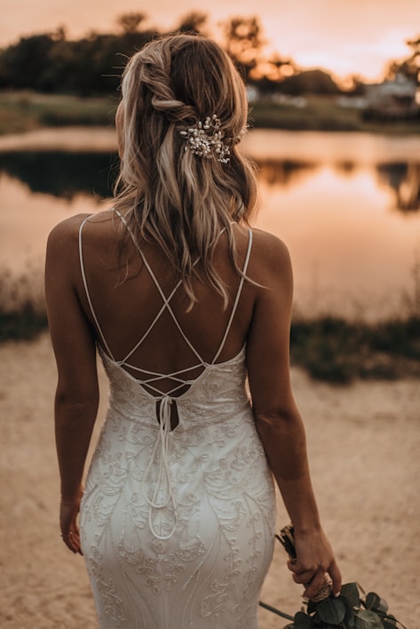 A woman in an elegant, white lace dress stands with her back to the camera, facing a serene, reflective body of water at sunset. Her blonde hair is styled in loose curls and adorned with an ornate hairpiece. She holds a small bouquet of flowers in one hand, complementing the romantic ambiance of the scene.