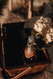 A bottle of champagne with a gold foil covering its cork is placed inside a wicker basket. Next to the bottle, there are white hydrangeas, creating an elegant and romantic setup. The lighting is warm and casts soft shadows, enhancing a cozy atmosphere.