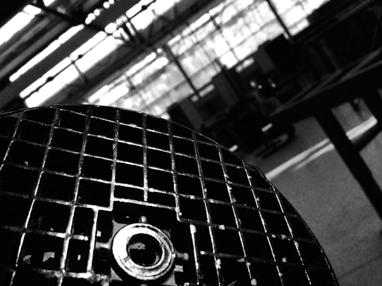 A black and white image featuring a close-up of a metallic grid or grate in the foreground, with a blurred industrial setting in the background. The grid has a circular knob or bolt in the center, suggesting machinery or equipment.