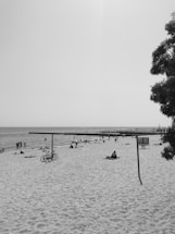 A sandy beach scene with people scattered across the shoreline, engaging in various leisure activities. Several individuals are walking along the water's edge, while others are lying on the sand, possibly sunbathing. Bicycles are parked under a simple canopy structure, and a tree is visible on the right side. The sky appears clear, and a long pier or jetty extends into the sea in the distance.