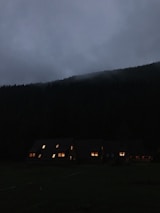 A serene, dark evening scene with a row of cabins nestled at the edge of a forested hillside. The sky is overcast with gray clouds, and soft glows of light are emanating from the windows of the cabins.