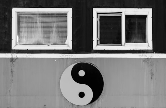 A yin-yang symbol is displayed prominently against a backdrop that includes two windows with white frames. The black and white color scheme emphasizes contrast, drawing focus to the symbol beneath the paired windows.