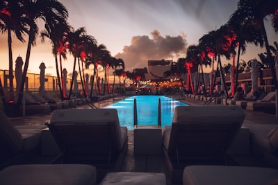 A luxurious poolside scene at sunset with rows of palm trees illuminated by red lights. The swimming pool glows with a vibrant blue, flanked by lounge chairs and cabanas. The sky is filled with dramatic clouds, and the cityscape is visible in the background, enhancing the ambiance.