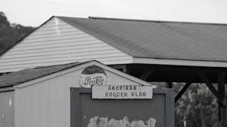 A small white structure with a triangular roof features an old Pepsi-Cola sign and a weathered sign reading 'American Soccer Club.' In the background, there are trees and a wooden pavilion with a gabled roof.