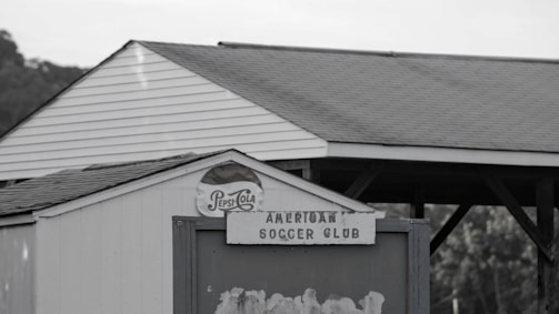 A small white structure with a triangular roof features an old Pepsi-Cola sign and a weathered sign reading 'American Soccer Club.' In the background, there are trees and a wooden pavilion with a gabled roof.