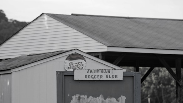 A small white structure with a triangular roof features an old Pepsi-Cola sign and a weathered sign reading 'American Soccer Club.' In the background, there are trees and a wooden pavilion with a gabled roof.