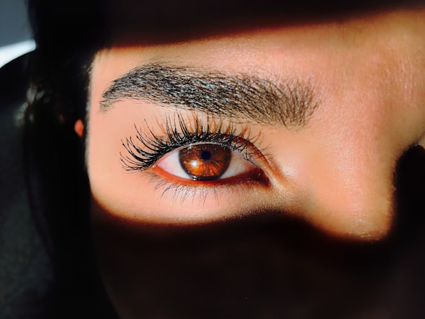 A close-up view of a human eye with long eyelashes, surrounded by shadow. The eye is amber-colored and bright, with light reflecting off its surface. The eyebrows are well-defined and dark, adding contrast to the image.