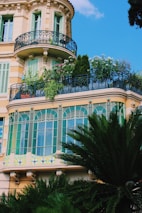 A building facade featuring ornate balconies with wrought iron railings and a lush rooftop garden. The windows are adorned with decorative green and blue detailing, and tall green plants are visible in the foreground.