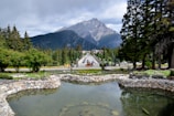 A serene landscape featuring a tranquil pond with clear water in the foreground, bordered by a stone wall. Behind the pond, a road lined with trees leads into a small town nestled at the foothills of a majestic mountain range. Tall pine trees and lush greenery surround the area, creating a harmonious blend between nature and civilization.