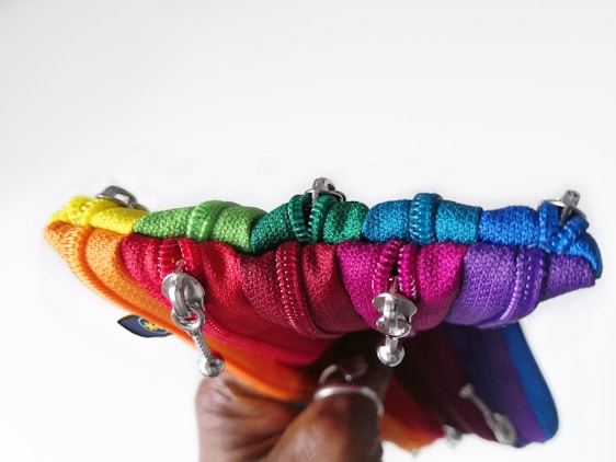 A collection of colorful zippered pouches, each in a different vibrant hue, is held together. The pouches are arranged in a rainbow sequence with visible zippers and metallic pull tabs, and are being held in a hand with a dark skin tone.