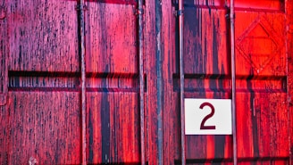 A close-up of a textured, painted metal surface with a mix of red and black vertical streaks. A white square with the number 2 is affixed to the surface.
