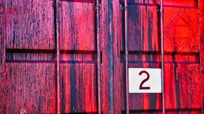 A close-up of a textured, painted metal surface with a mix of red and black vertical streaks. A white square with the number 2 is affixed to the surface.