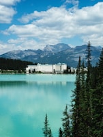A grand hotel sits by the edge of a tranquil, turquoise lake, surrounded by lush evergreen trees. Towering mountains form an impressive backdrop under a partly cloudy, blue sky.