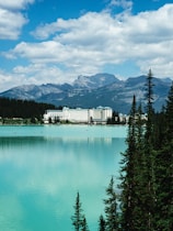 A grand hotel sits by the edge of a tranquil, turquoise lake, surrounded by lush evergreen trees. Towering mountains form an impressive backdrop under a partly cloudy, blue sky.