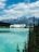 A grand hotel sits by the edge of a tranquil, turquoise lake, surrounded by lush evergreen trees. Towering mountains form an impressive backdrop under a partly cloudy, blue sky.