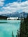 A grand hotel sits by the edge of a tranquil, turquoise lake, surrounded by lush evergreen trees. Towering mountains form an impressive backdrop under a partly cloudy, blue sky.