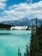 A grand hotel sits by the edge of a tranquil, turquoise lake, surrounded by lush evergreen trees. Towering mountains form an impressive backdrop under a partly cloudy, blue sky.