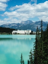A grand hotel sits by the edge of a tranquil, turquoise lake, surrounded by lush evergreen trees. Towering mountains form an impressive backdrop under a partly cloudy, blue sky.