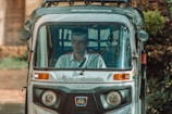 A person is seated inside a motorized rickshaw, often referred to as a tuk-tuk. The front view shows the driver behind the glass windshield, with a rural road and blurred greenery in the background. The rickshaw is marked with branding on the front.