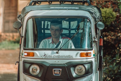 A person is seated inside a motorized rickshaw, often referred to as a tuk-tuk. The front view shows the driver behind the glass windshield, with a rural road and blurred greenery in the background. The rickshaw is marked with branding on the front.