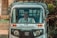 A person is seated inside a motorized rickshaw, often referred to as a tuk-tuk. The front view shows the driver behind the glass windshield, with a rural road and blurred greenery in the background. The rickshaw is marked with branding on the front.