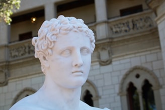 A marble statue of a male figure with curly hair is prominently displayed in the foreground, set against the backdrop of an ornate building with stone balconies and arched windows. The lighting emphasizes the smooth texture of the statue's surface.