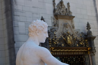 A white marble statue of a male figure is positioned in front of an ornate building. The architecture features intricate carvings, including a crest and decorative gold elements. The statue is depicted from a side angle, showcasing detailed curls in the hair.