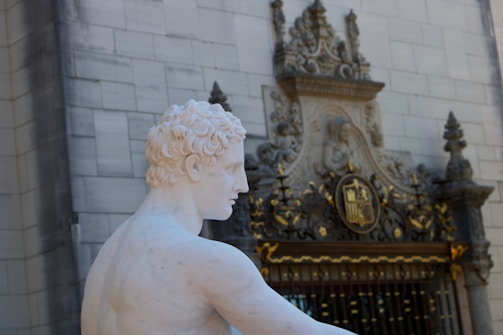 A white marble statue of a male figure is positioned in front of an ornate building. The architecture features intricate carvings, including a crest and decorative gold elements. The statue is depicted from a side angle, showcasing detailed curls in the hair.