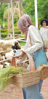 A person wearing a headscarf and casual clothing is holding a wicker basket filled with grass and a plastic bottle, feeding sheep in a sunny outdoor setting. There is another person in the background, wearing a hat. The setting appears to be a farm or petting zoo with greenery and rustic architecture.